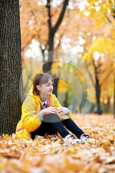 Sad teen girl sits near tree in autumn park. Bright yellow leaves and trees