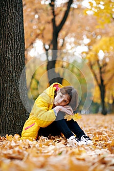 Sad teen girl sits near tree in autumn park. Bright yellow leaves and trees