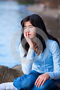 Sad teen girl sitting on rocks along lake shore, lonely expression