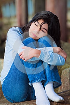 Sad teen girl sitting on rocks along lake shore, lonely expression