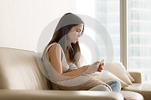 Sad teen girl using phone while sitting on sofa