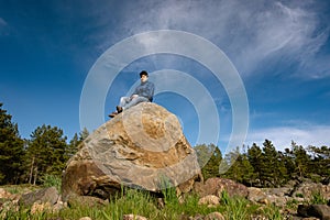 Sad teenage boy sits at top of big rock, pine trees and blue sky at background