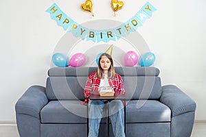 A sad teenage girl sits alone at home with a cake in a room decorated in honor of her birthday and crying, unhappy, copy space.