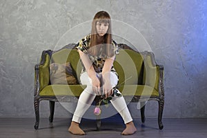 Sad teenage girl sits on green sofa. On gray background. Studio.