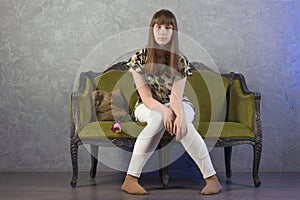 Sad teenage girl sits on green sofa. On gray background. Studio.