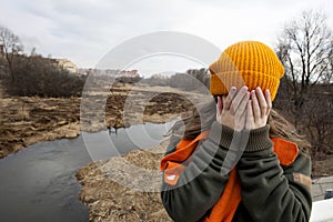 Sad teenager in orange knitten hat and scarf closed her face by hands