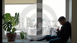 Sad teenager sitting and looking out of the window and using mobile cell phone for social media, watch video at home
