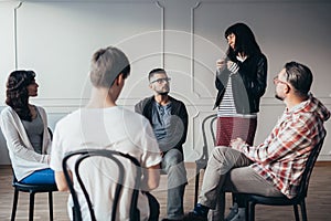 Sad teenager stands in front of a group of people of different ages during a therapy group meeting and talks about his problems