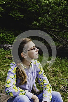 Sad thoughtful teen girl with glasses sitting in the forest on the nature alone.
