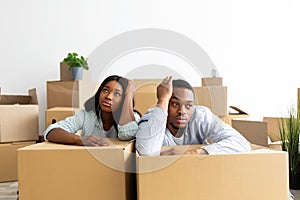 Sad and tired black couple leaning on unpacked boxes, sitting in the room full of boxes, ready to move to new house