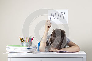 Sad tired frustrated boy sitting at the table with many books and holding paper with word Help. Learning difficulties