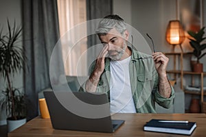 Sad tired retired caucasian man with beard takes off his glasses, rubs eyes looks at computer in room interior