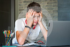 Sad tired teenager in white shirt sitting behind desk in kitchen  near laptop and study. Depressed Boy in earphones makes homework