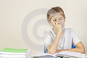 Sad tired upset schoolboy covering face with his hands sitting at table with pile of school books and notebooks at home