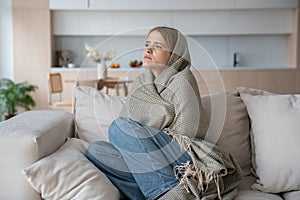 Sad tired woman sitting on couch at home wrapped in plaid in living room. Flu season.
