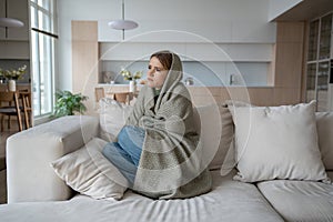 Sad tired woman sitting on couch at home wrapped in plaid in living room. Flu season.