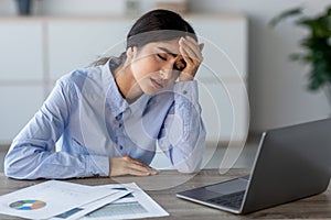 Sad tired young indian lady employee presses hand to head at workplace with laptop in office or home photo