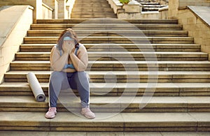 Sad tired young overweight woman with sports mat sitting on stone steps and crying