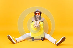 Sad traveler tourist woman in summer casual clothes, hat sit near suitcase on yellow orange background. Female photo