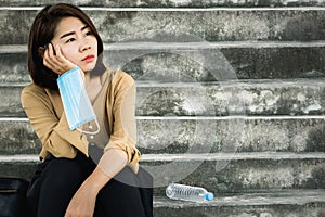 Sad unemployed Asian business woman hand holding protective mask sitting alone on stairs thinking of future, covid 19 crisis