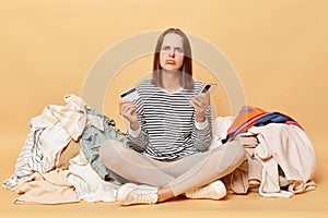 Sad unhappy Caucasian woman posing near heap of multicolored unsorted clothes isolated over beige background looking at camera