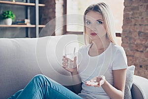 Sad unhappy depressed woman is holding a pill in a hand and a glass of water, she is very ill and stayed at home instead of going