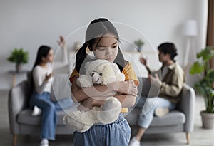 Sad unhappy despaired korean teenage girl hugs toy, parents quarrel and shout in living room interior, blurred