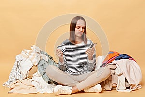Sad unhappy disappointed Caucasian woman posing near heap of multicolored unsorted clothes isolated over beige background has not
