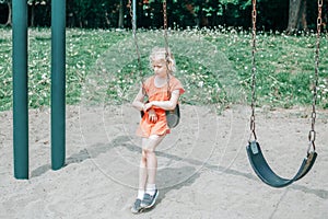 Sad unhappy girl is swinging on swing set in park outdoor alone. Upset bored kid waiting for friend on playground. Lonely child is