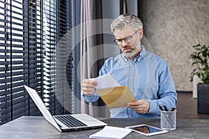 Sad unhappy man sitting at workplace inside office, senior mature gray-haired businessman taking mail letter, envelope