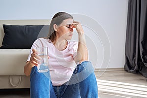 Sad unhappy mature woman sitting at home on floor with glass of water