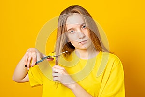 Sad and unhappy teen girl cutting her hair with scissors while standing over yellow background.
