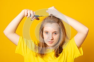 Sad and unhappy teen girl cutting her hair with scissors while standing over yellow background. Young student experiments with her