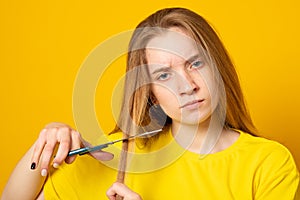 Sad and unhappy teen girl cutting her hair with scissors while standing. Young student experiments with her hairstyle at home