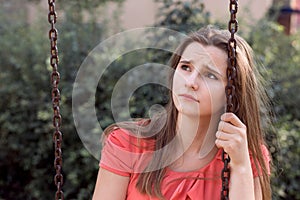 Sad unhappy teen girl with long brunette hair sitting on a swing sadly looking up