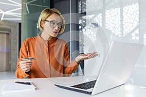 Sad and unhappy woman inside workplace office with laptop holding bank credit card, businesswoman cheated got negative