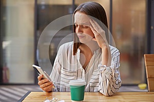 Sad unhappy woman wearing bluse sitting in cafe using mobile phone having troubles serfung internet having job problems frowning