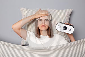 Sad unhappy woman in white T-shirt and sleeping eye mask lie in bed on pillow under blanket isolated on gray background making