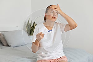 Sad unhappy young adult pregnant woman with pregnancy test posing in her bedroom, wearing white t shirt, looking away with sorrow