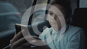 Sad, unhappy young boy riding in car through city during rainy day, using social network on his smartphone