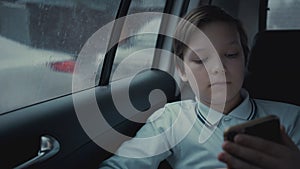 Sad, unhappy young boy riding in car through city during rainy day, using social network on his smartphone