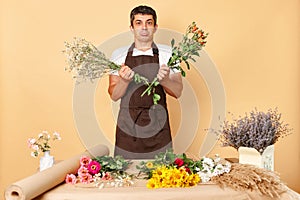 Sad unhappy young male in brown apron working in flower shop standing isolated over beige background holding two bouquets looking