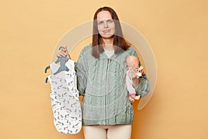 Sad unhappy young pregnant woman with baby bodysuits isolated over beige background holding doll toy looking at camera with upset