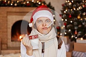 Sad unhealthy woman wearing santa claus hat, being wrapped in warm scarf, sitting near Christmas tree, measuring temperature with