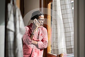 Sad upset African man having difficult conversation on phone, standing by window at home