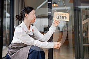 A sad and upset Asian female coffee shop owner is hanging a close sign at the entrance door