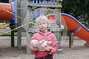 Sad upset Caucasian girl with baby toy on closed playground outdoor. Kids play area locked with yellow caution tape in Toronto,