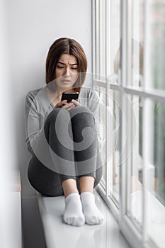 Sad upset caucasian millennial female sitting on windowsill at home and reading message on phone with bad news