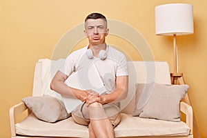 Sad upset man wearing white T-shirt and shorts sitting on sofa against beige background holding closed laptop looking at camera