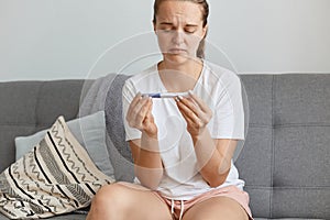 Sad upset young adult woman with sorrow feelings wearing white t shirt holding pregnancy test in hands with positive result, being